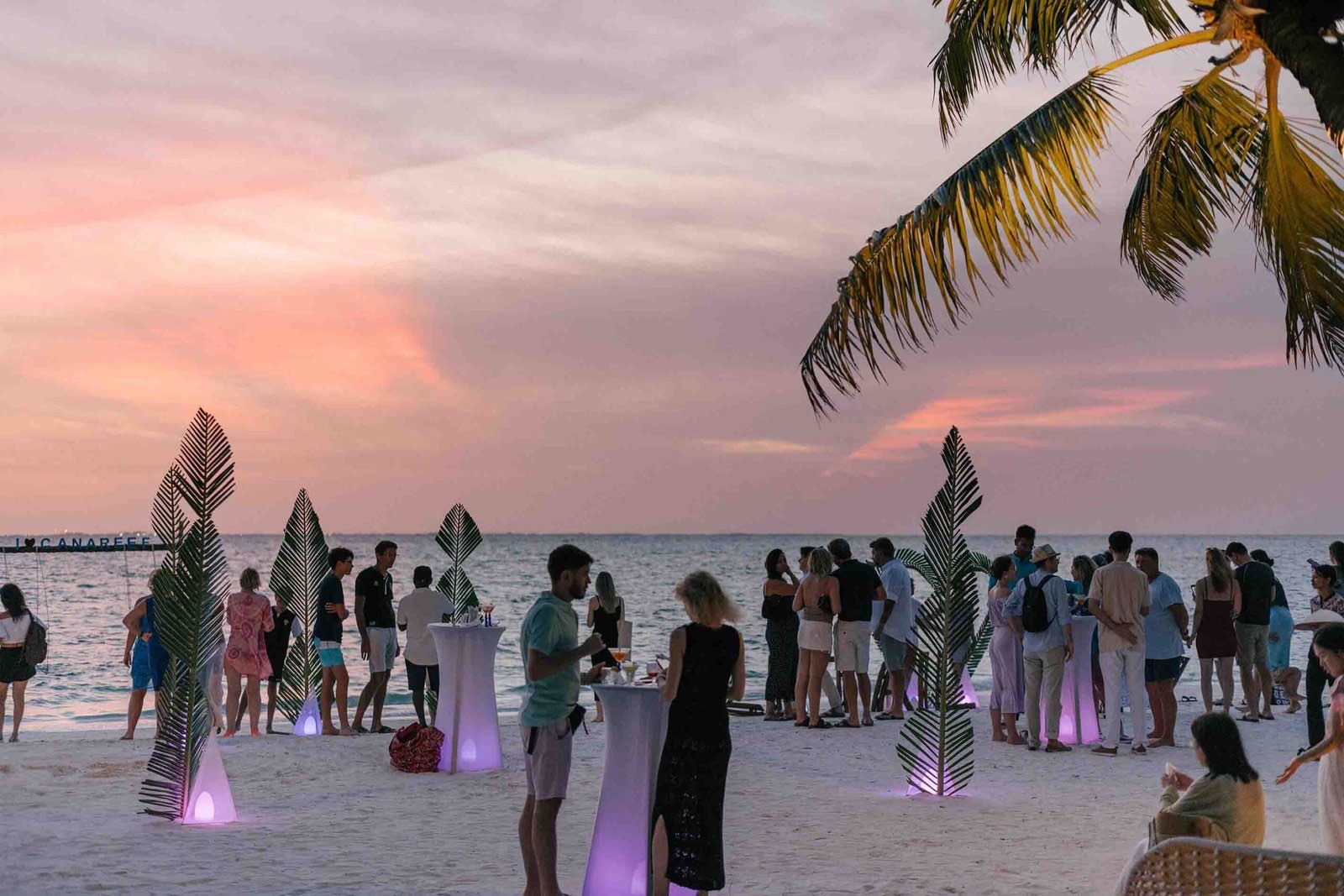An evening beach reception at sunset: guests with cocktails around lit tables on the sand under a pink sky.