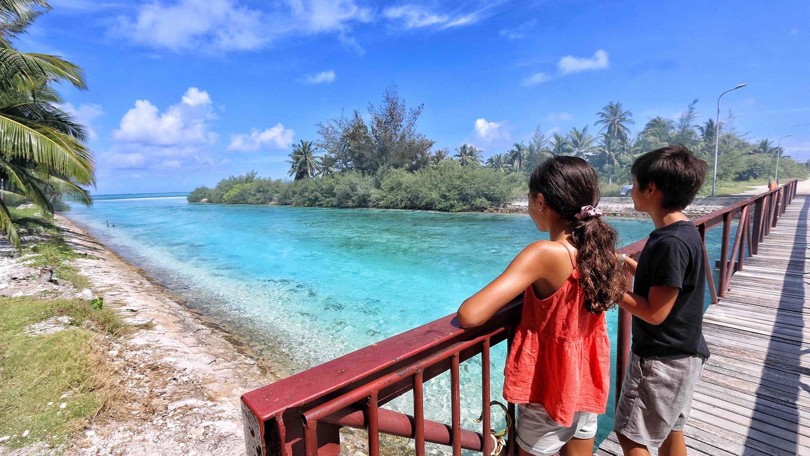 Two young people leaning on a red bridge railing, looking across bright turquoise water towards a palm-covered local island.