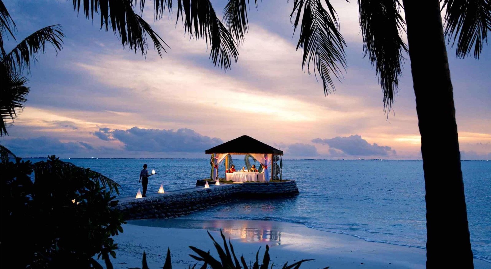 A dinner table dressed on the sand at the water's edge, lit for the evening.